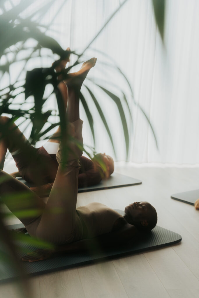 Vrouw ligt op mat met benen verticaal gestrekt achter grote groene plant, natuurlijk daglicht valt door raam, serene sfeer in pilates studio met focus op ademhaling en verlenging.