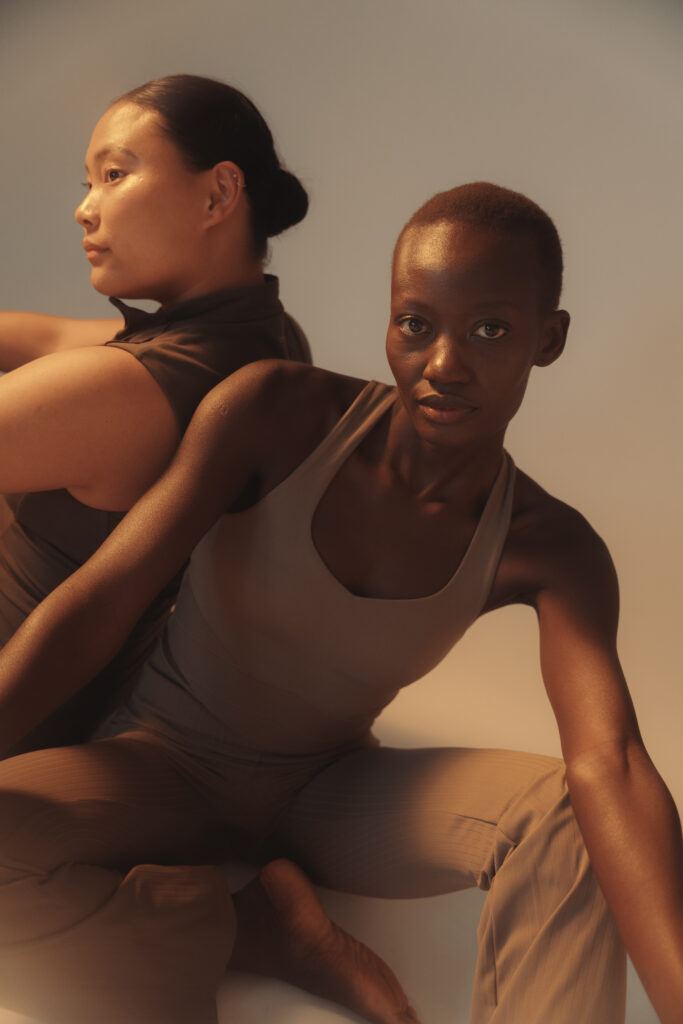 Twee vrouwen zitten naast elkaar in pilates outfit, kalme poses, warm licht creëert zachte schaduwen, beeld van verbinding en samen zijn in de studio.