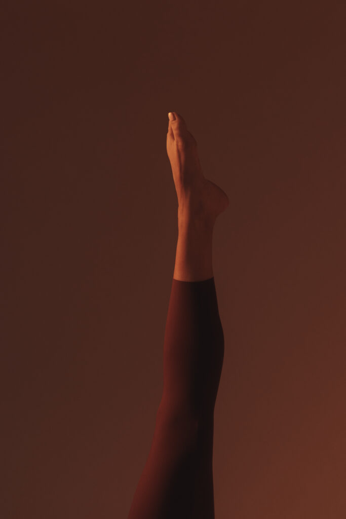 Gestrekt been verticaal omhoog tegen donkere achtergrond met warme oranje verlichting, minimalistische compositie met focus op lijn en beweging tijdens pilates practice.